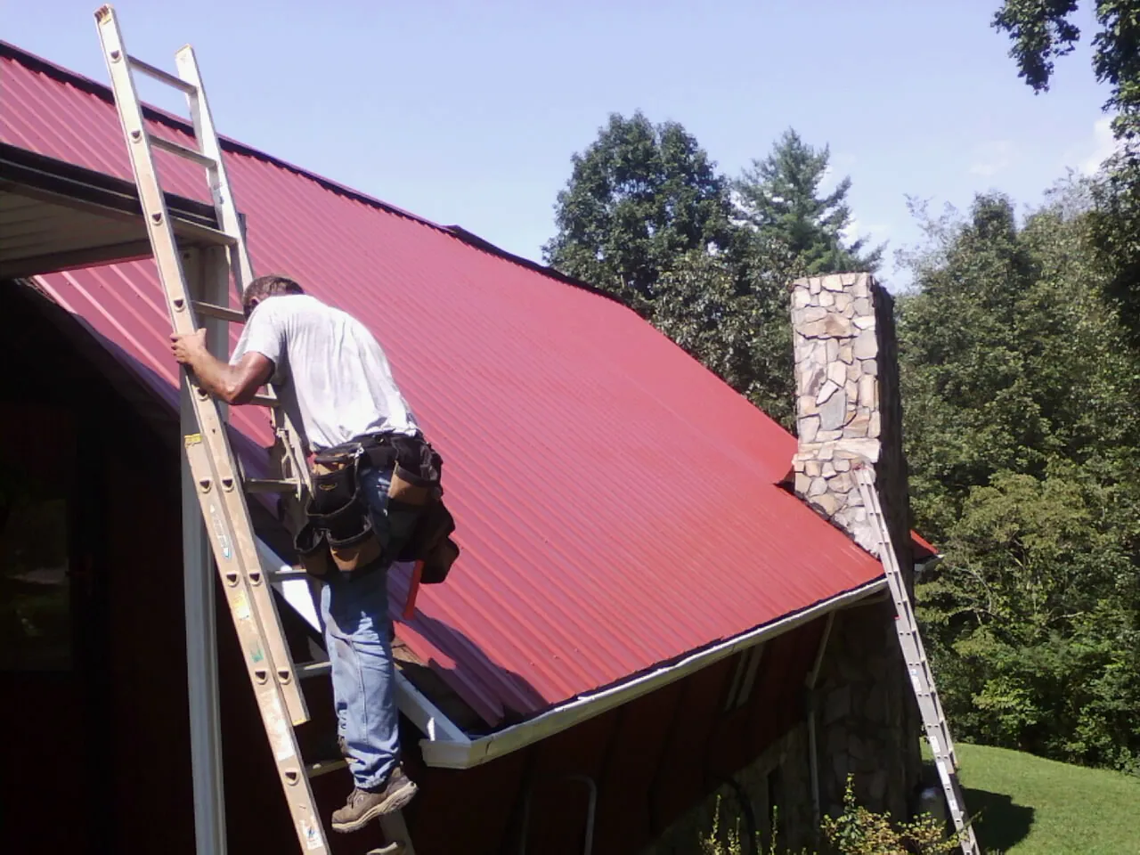 Roofer on a ladder beside a red metal roof and stone chimney — Soffit & Fascia Repair by Roofing Brothers in Lusk